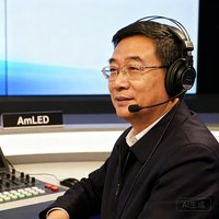 浙江电视台演播技术部主任王建先生半身照，头戴专业耳机，坐在演播控制台前，身后是AmLED背景屏